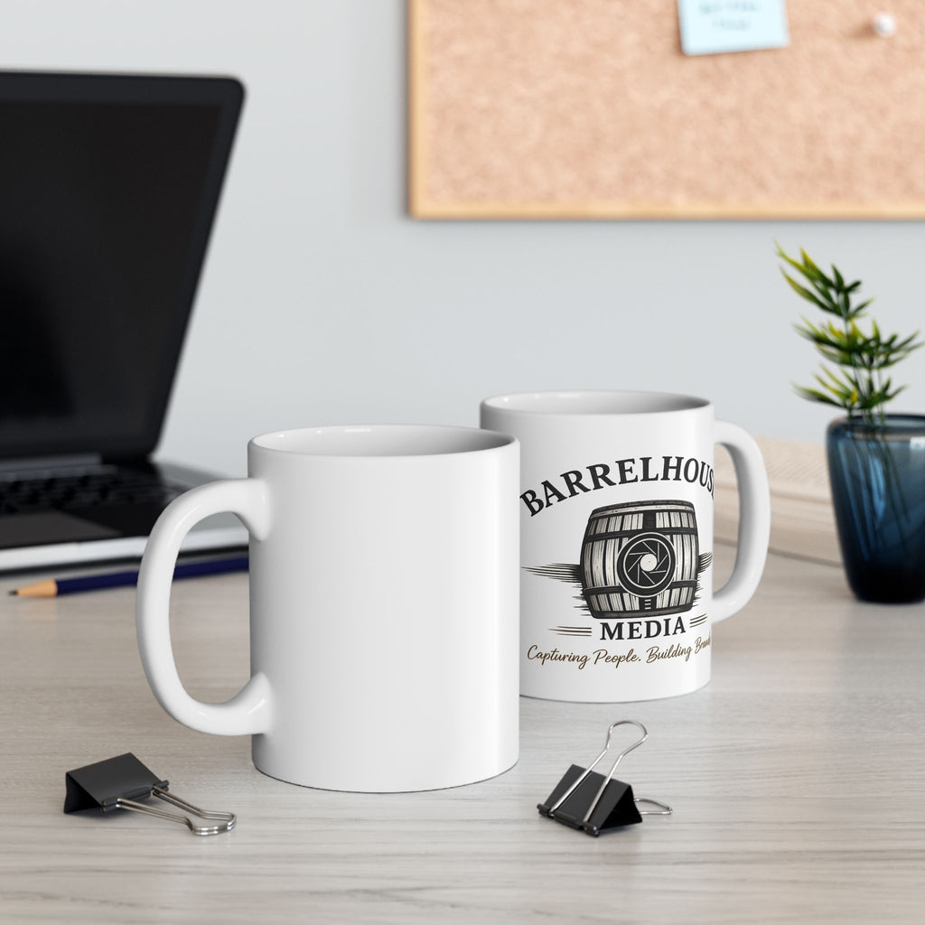 Two white ceramic Barrelhouse Media coffee mugs, one with logo, on a light wood desk with laptop and plant.