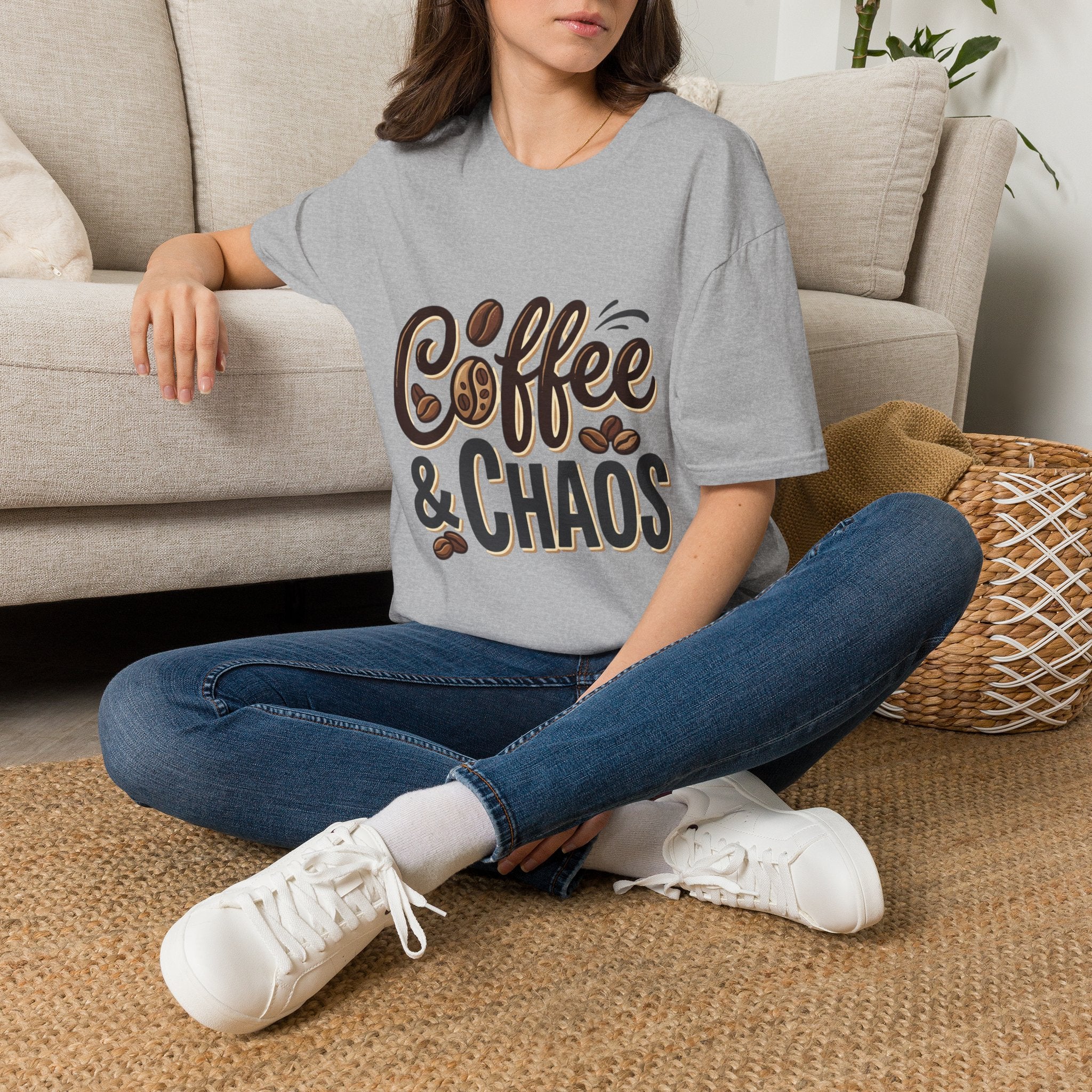 Woman wearing a gray "Coffee & Chaos" t-shirt, blue jeans, and white sneakers, sitting on a rug.