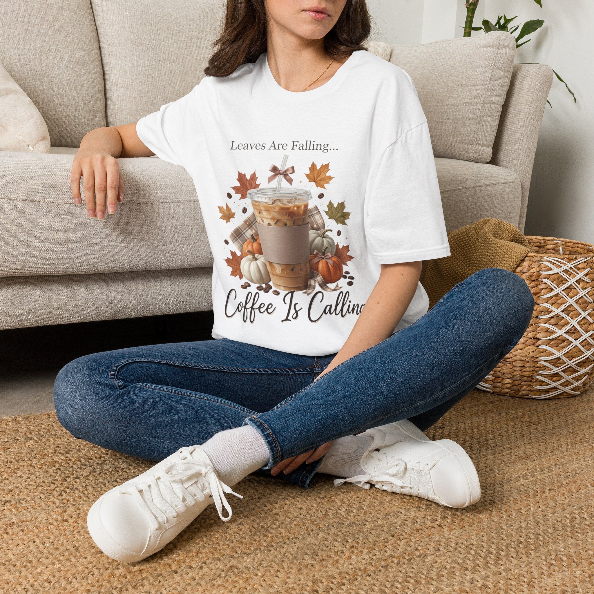 Front view of a white "Leaves Are Falling... Coffee Is Calling" t-shirt with an iced latte, leaves, and pumpkins graphic, worn by a woman.