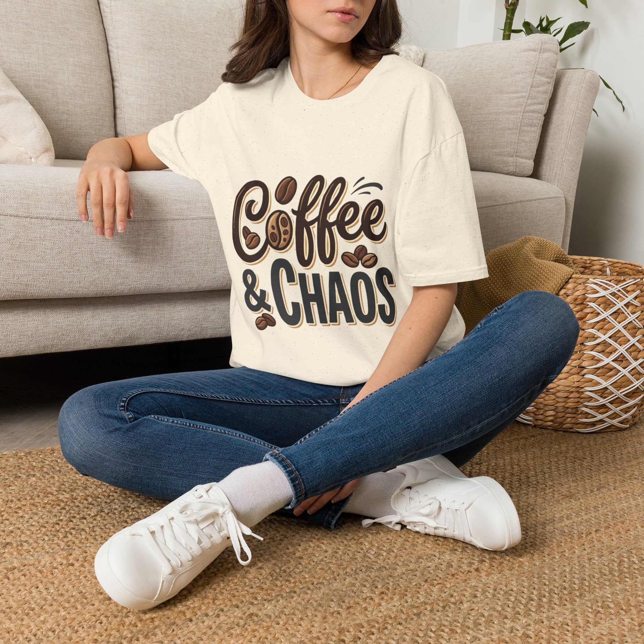 Front view of a woman wearing a white "Coffee & Chaos" t-shirt, blue jeans, and white sneakers, sitting on a rug.