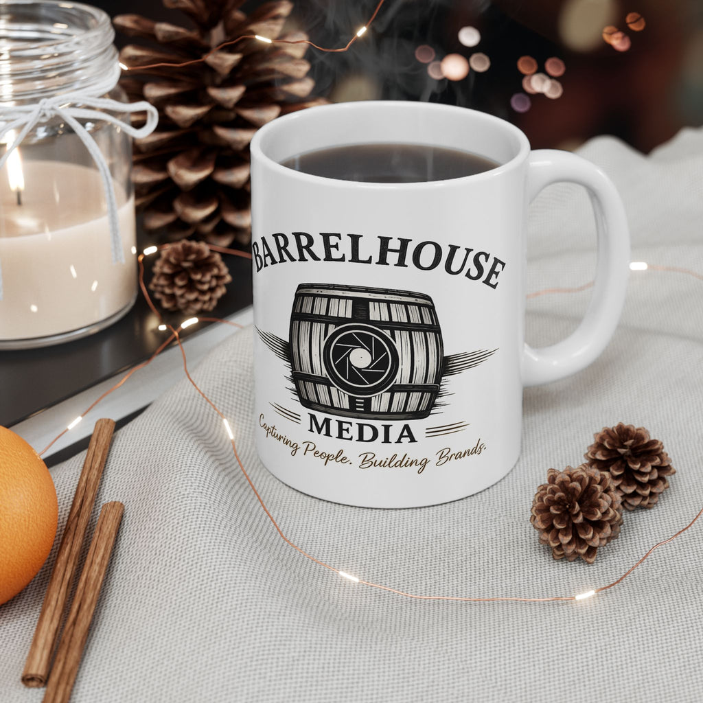 Front view of a white ceramic Barrelhouse Media logo coffee mug with a barrel and camera lens graphic, filled with dark liquid, surrounded by pinecones, cinnamon sticks, and a lit candle.